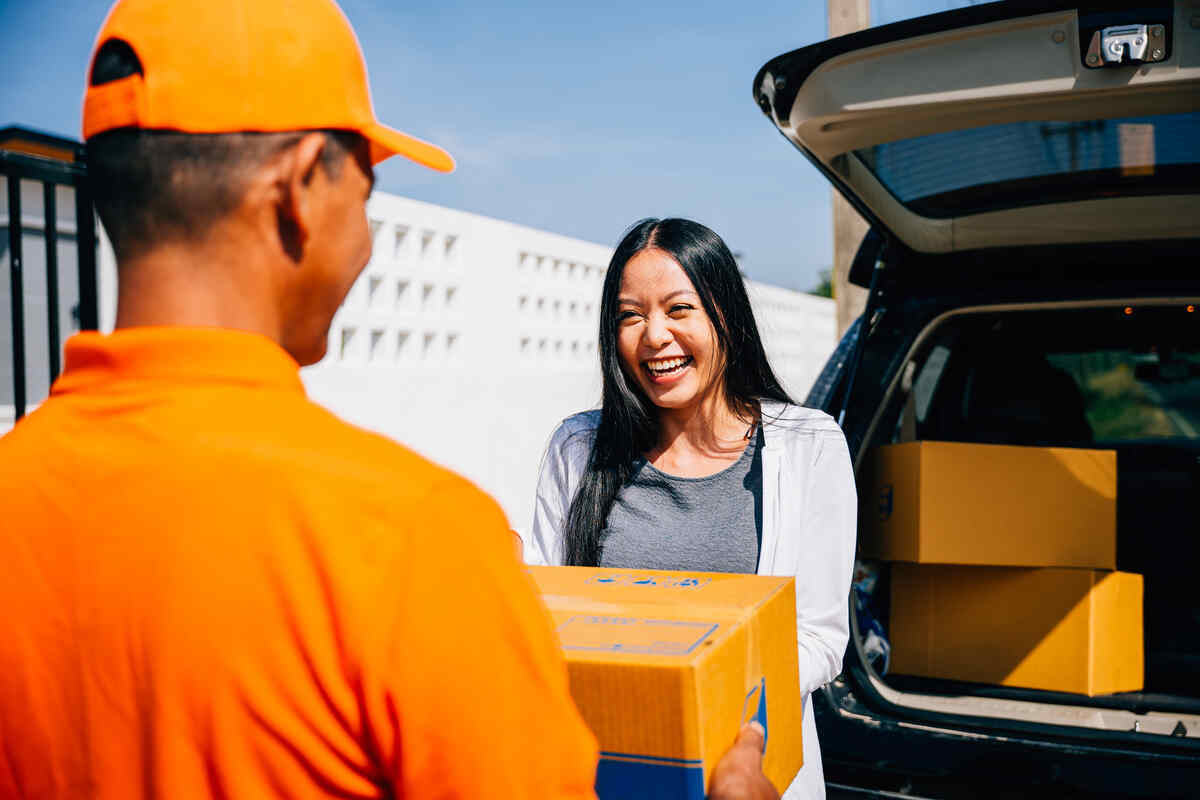 Smiling woman receives a package from a delivery driver beside an open car trunk on a sunny day.