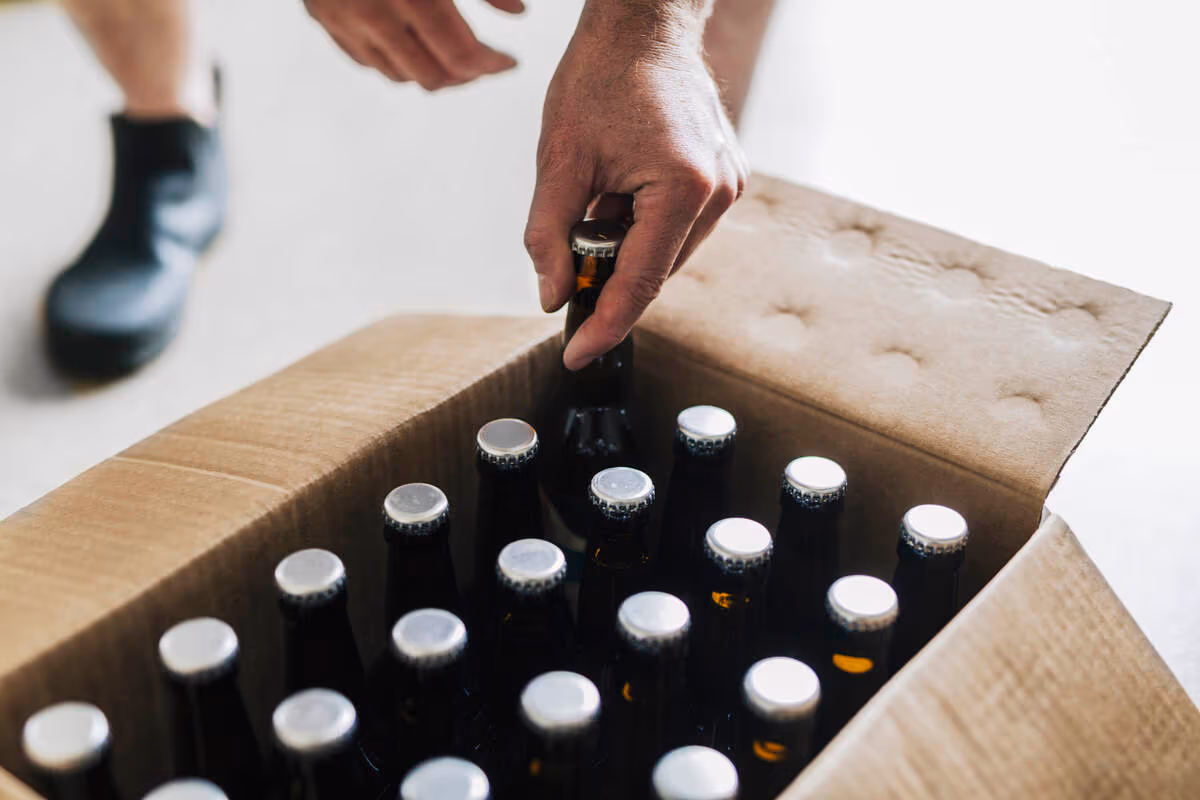 A person's hand is reaching into a cardboard box filled with amber beer bottles sealed with silver caps. Partial view of legs and shoes is visible nearby.