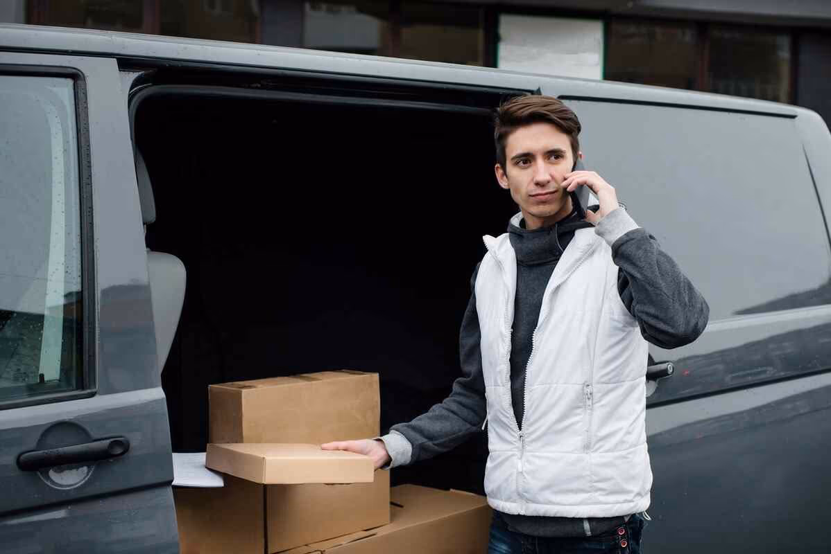 Person in white vest stands by an open van, holding a cardboard box and on the phone, focused on deliveries.