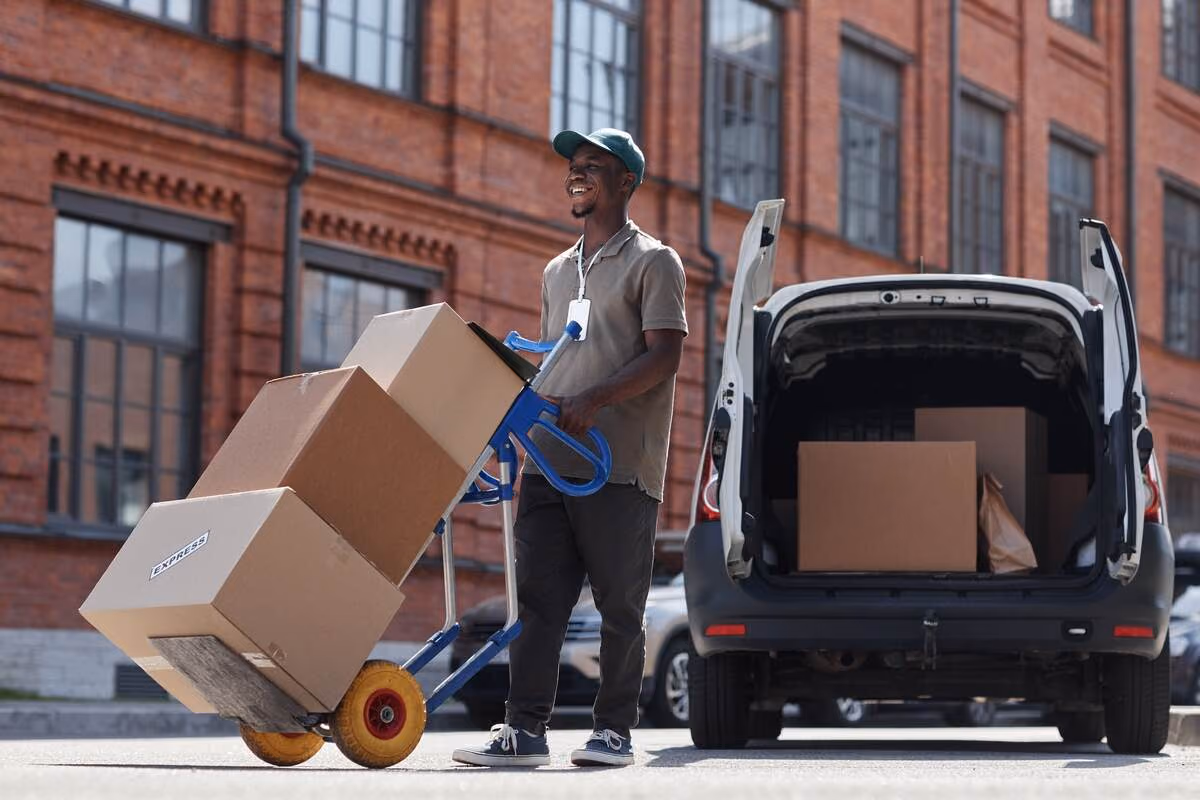 Smiling delivery worker moves boxes on a hand truck near a parcel van.