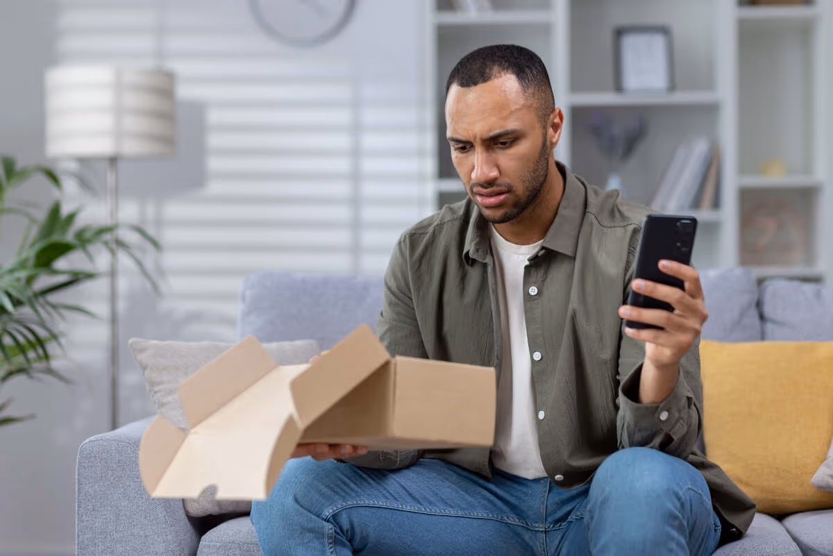 Worried man on couch holding empty box, looking at smartphone in cozy living room.